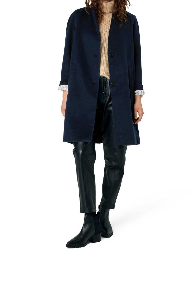 Femme portant un manteau bleu marine, un pull en tricot beige, un pantalon en cuir noir et des bottines noires, debout sur un fond blanc.