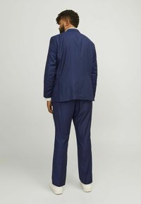 Costume bleu marine avec une coupe ajustée. La veste présente une fente unique et une texture lisse, associée à un pantalon assorti et des chaussures blanches.
