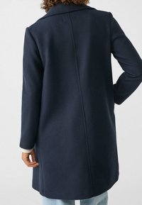 Mørkeblå uldfrakke med skræddersyet design, enkeltbælte foran og lange ærmer. Har et lige snit og diskrete syningsdetaljer.