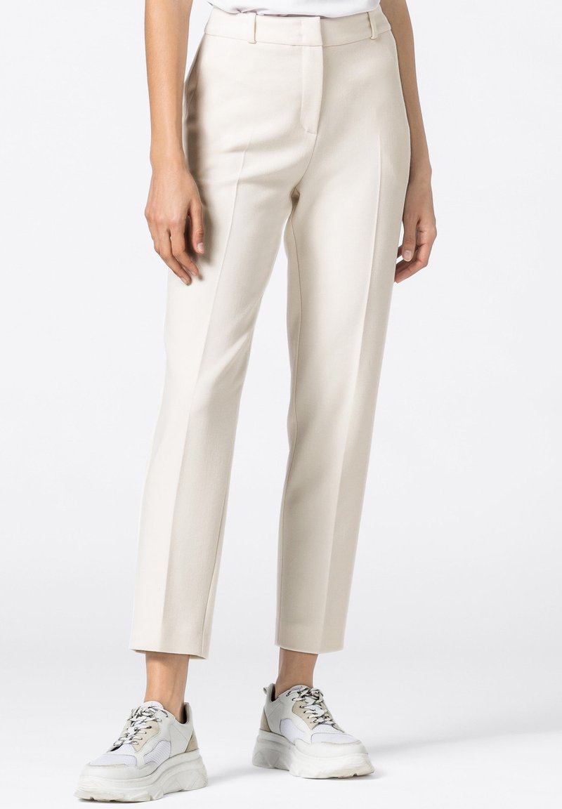 Femme portant un pantalon beige taille haute sur-mesure associé à des baskets blanches épaisses, debout sur un fond blanc.