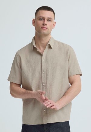 Nuori mies, jolla on lyhyet hiukset ja joka pukeutuu beigeen, lyhythihaiseen nappipaitaan, pitää käsiään ristissä vaaleanharmaan taustan edessä.