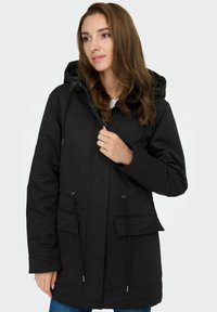 Črna parkas jakna s kapuco, z vrvico na pasu in dvema sprednjima žepoma. Narejena iz gladkega materiala z oprijetim dizajnom.