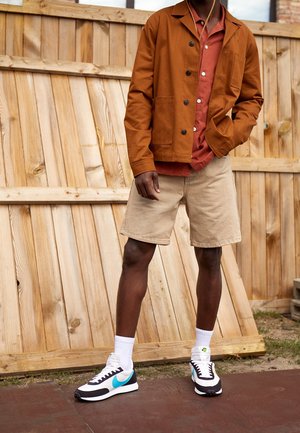 Brown cotton jacket, red button-up shirt, beige shorts. Black and white sneakers with teal accent. Wooden fence background.