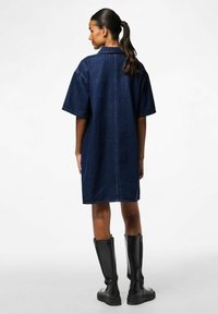 Tumši zila džinsu kleita ar īsām piedurknēm, funkcionālu apkakli un kontrastējošu šuvumu. Apvienota ar melniem zābakiem līdz ceļiem.