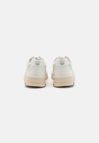 White sneakers with textured sides, beige rubber soles, and a subtle logo on the heel. Laces are untied with a soft textile mesh upper.