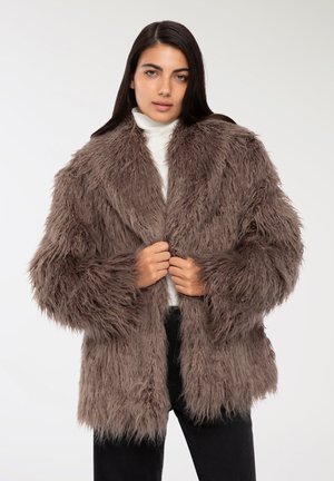 Cappotto in pelliccia sintetica marrone con texture lunga e shaggy, colletto ampio e apertura frontale. Indossato sopra un dolcevita bianco e pantaloni neri.