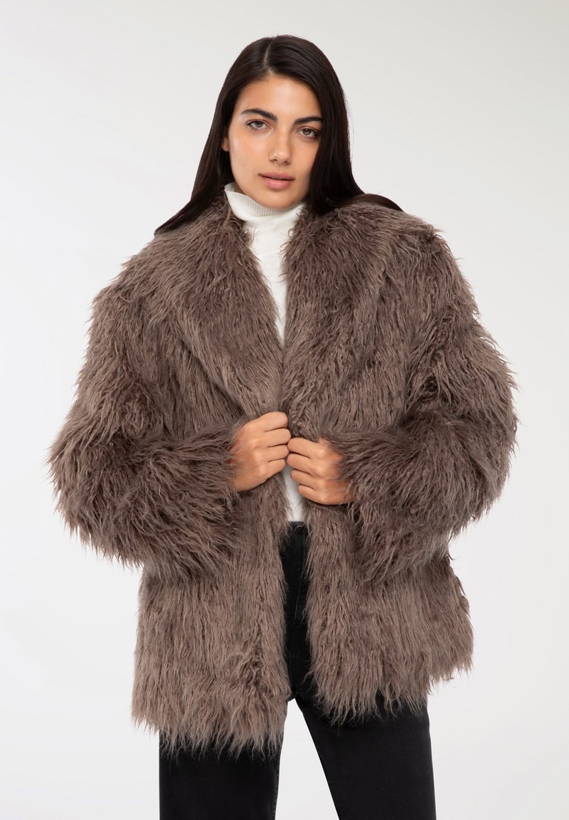 Cappotto in pelliccia sintetica marrone con texture lunga e shaggy, colletto ampio e apertura frontale. Indossato sopra un dolcevita bianco e pantaloni neri.