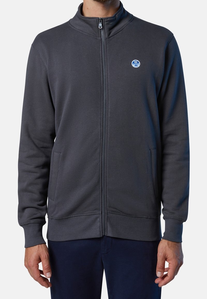 Homme portant une veste zippée gris foncé avec un petit logo rond sur le côté gauche de la poitrine et deux poches avant, debout les mains détendues.
