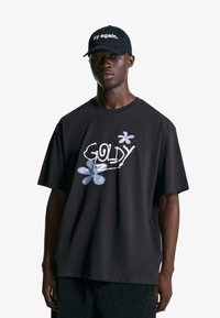 Schwarzes T-Shirt mit dem Text "GOLDY" und blauen Blumenmotiven. Getragen mit einer schwarzen Mütze, die den Text "try again" trägt. Lässiges Design, entspannte Passform.