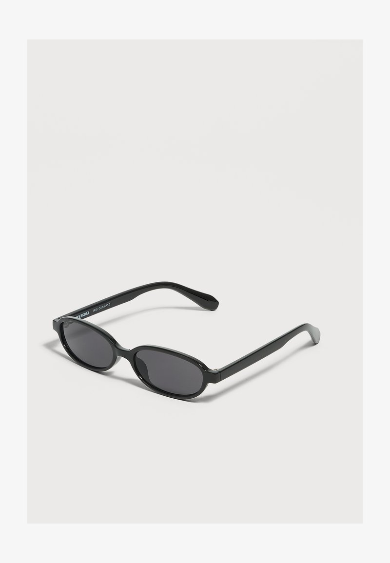 Lunettes de soleil ovales noires avec des verres foncés et des montures fines posées sur une surface claire.