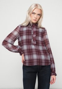 Noor naine, kellel on pikad blondid juuksed, kannab õhukest pikavarrukatega burgundi ja valge ruudulist pluusi ning tumedaid teksaseid, poseerides lihtsa tausta ees.