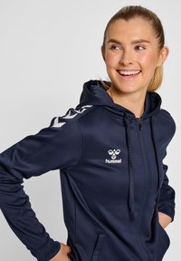 Felpa con zip di colore blu navy, con una superficie testurizzata, caratterizzata da un logo bianco e accenti a chevron sulle maniche, e cappuccio con cordino. Vestibilità standard.