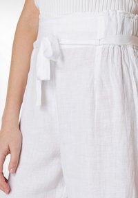 Pantaloni bianchi in linen con vita alta, tasche laterali e una cintura con dettaglio a fiocco. Il tessuto ha una texture liscia e leggera.