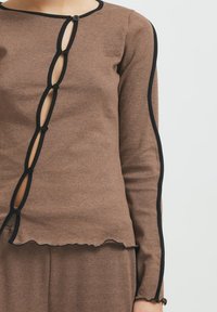 Brown long-sleeve top with black trim and button closure, featuring a unique cut-out design along the front and scalloped hem.