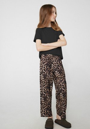 Mädchen steht mit verschränkten Armen, trägt ein schwarzes kurzärmeliges Shirt, weit geschnittene Hose mit Leopardenmuster und dunkle Slip-On-Schuhe auf einfachem Hintergrund.