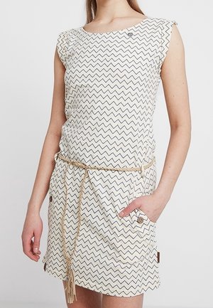 Femme portant une robe crème sans manches, longueur genoux, avec un motif en zigzag noir et une fine ceinture tressée, main dans la poche côté droit.