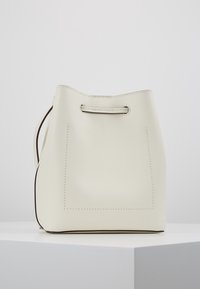 Krémová kožená taška ve tvaru kbelíku se stehujícími detaily a stahovací šňůrkou, umístěná na bílém podstavci proti jednoduchému pozadí.