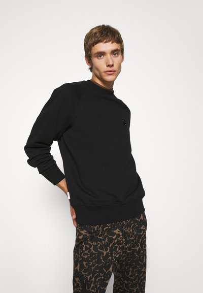 Sweat-shirt noir avec un col rond et des poignets côtelés, arborant un petit logo sur la poitrine. Porté avec un pantalon brun à motifs.