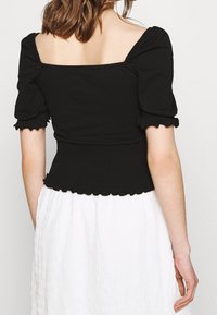 Black fitted top with short puff sleeves, square neckline, and smocked waist. Features ruffled cuffs. Paired with a white textured skirt.