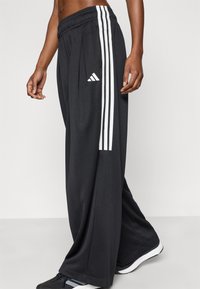 Μαύρο αθλητικό παντελόνι με ελαστική μέση, που διαθέτει τρεις λευκές κάθετες ρίγες στις πλευρές και λογότυπο της Adidas στο αριστερό μηρό.