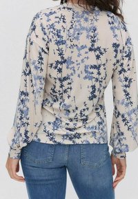 Langarmbluse in hellbeige mit blauem Blumenmuster. Weicher Stoff, lockere Passform und Raffung an den Bündchen. Kombiniert mit blauen Jeans.