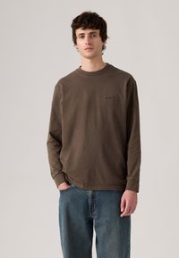 Tricou lung, maro, din bumbac moale, cu guler rotund, având un logo subtil pe piept și o croială lejeră, asociat cu blugi albaștri.