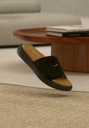 Sandale noire à glissière avec une large bride ornée d'une boucle carrée, une semelle intérieure couleur fauve et une semelle texturée, flottant au-dessus d'une surface de tapis tissé.