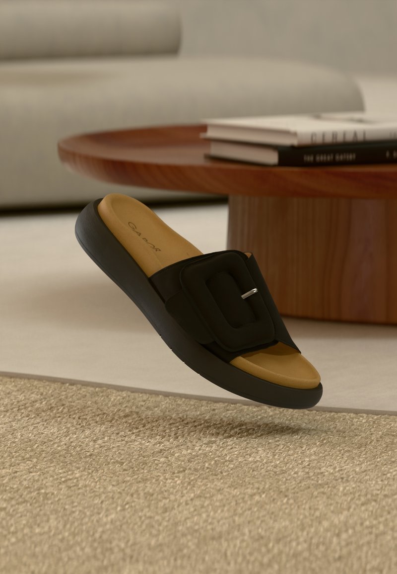 Μαύρο σανδάλι τύπου slide με φαρδιά λουρί που διαθέτει τετράγωνη αγκράφα, ταμπά πάτος και υφασμένη σόλα, που αιωρείται πάνω από υφαντό χαλί.