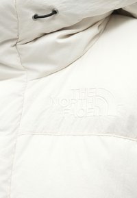 Doudoune blanche avec texture matelassée, logo brodé "THE NORTH FACE" et capuche réglable à cordon.