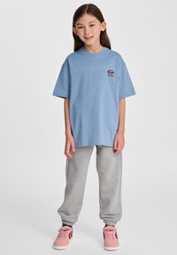 Niebieska bawełniana koszulka z okrągłym dekoltem i krótkimi rękawami, w zestawie z szarymi dresami i różowymi trampkami. Na piersi znajduje się logo.