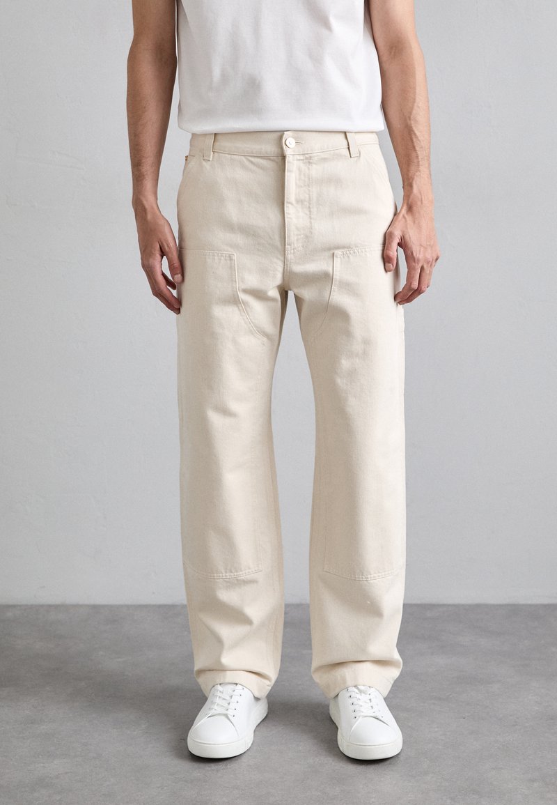 Beige cotton trousers with a relaxed fit, featuring front pockets, a button closure, and raw edge detailing, paired with white sneakers.