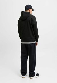 Schwarzer Kapuzenpullover, lockere Passform, kombiniert mit lässigen blauen Hosen. Modell trägt dunkle Sneakers mit weißen Akzenten, von hinten gesehen.