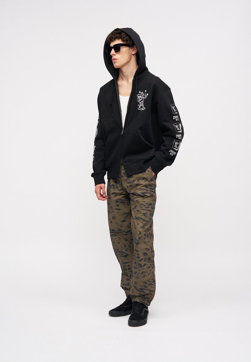 Junger Mann trägt schwarzen Hoodie mit weißem Motiv, camouflagemusterte Jogginghose, schwarze Sneakers, Sonnenbrille, steht mit einer Hand in der Hosentasche.