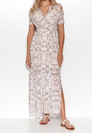 Femme portant une robe portefeuille beige à motifs, manches courtes et fente latérale, assortie à des sandales plates blanches sur un fond uni.