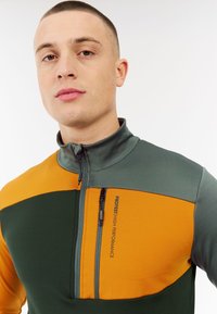 Fleece pullover met hoge kraag; heeft kleurblokken in groen, oranje en grijs; inclusief een zippered borstzak en logo detail.