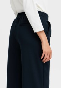 Pantaloni larghi blu navy con vita elasticizzata, dotati di tasche laterali e di un tessuto liscio e strutturato. Sopra, una maglia bianca a maniche lunghe.
