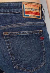 Mörkblå denimjeans med en synlig märkesetikett, kontrasterande orange sömmar och en liten röd accent på bakfickan.