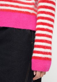 Kootud triibuline kampsun, millel on punased ja valged triibud ning erkroosa soonikserv. Kandmisega tumesiniste teksadega, mis näitavad tihedat istuvust.