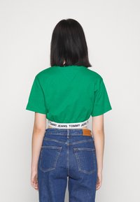 Zelené zkrácené tričko s kulatým výstřihem, spárované s modrými džíny s vysokým pasem, které mají značkový pas a koženou nášivku na zadní straně.