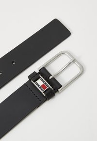 Zwarte leren riem met een zilveren rechthoekige gesp. De gesp heeft een logo met rode, witte en blauwe details. Soepele textuur, minimalistisch ontwerp.