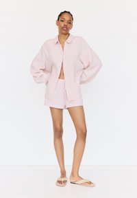 Lichtroze, oversized, overhemd met knopen en lange mouwen van textuurstof, gecombineerd met bijpassende shorts en beige sandalen.