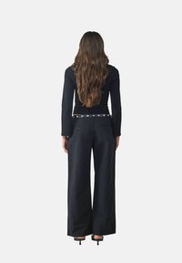 Zwarte langarmige top in combinatie met wijde zwarte broek. De broek heeft zilveren hardware-accenten en zakken. Het haar is lang en golvend.