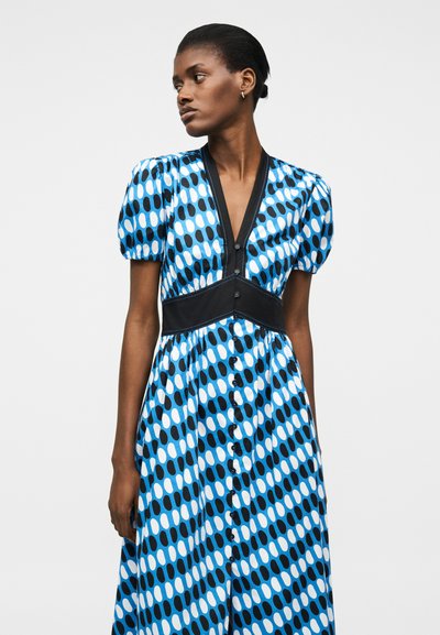 Γυναίκα που φοράει φόρεμα με μοτίβο σε μπλε, μαύρο και λευκό, με φουσκωτά μανίκια και V-λαιμόκοψη, στέκεται μπροστά σε απλό λευκό φόντο.