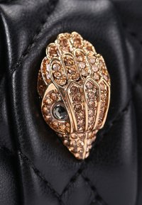 La pelle trapuntata nera presenta una spilla a forma di conchiglia in tono oro, adornata con piccoli cristalli marrone chiaro e una gemma centrale scura.