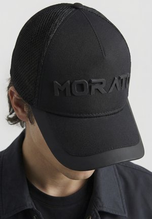 Personne portant une casquette noire à dos en maille avec l'inscription "MORATO" en relief à l'avant, associée à une chemise foncée et un t-shirt.