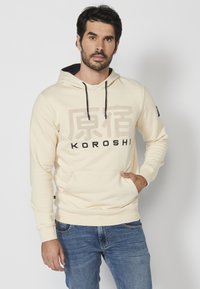 Sudadera crema con detalles en negro, que incluye un bolsillo frontal estilo canguro, capucha con cordón y un texto destacado "KOROSHI" en negro y beige.