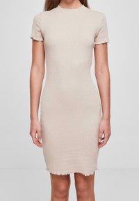 Robe courte ajustée de couleur beige clair à manches courtes avec des bords festonnés aux manches et à l'ourlet, portée par une personne debout sur fond uni.