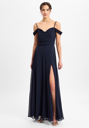 Navyblaues Abendkleid aus weichem Stoff, mit gerafftem Oberteil, schulterfreien Ärmeln und einem Oberschenkel-spitzem Schlitz. Silberne Sandalen vervollständigen den Look.