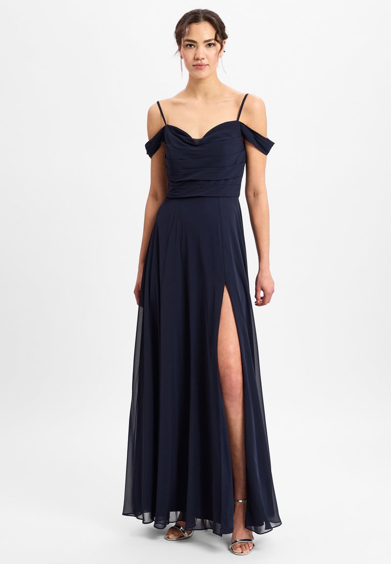 Navyblauwe avondjurk van zachte stof, met een gerimpelde lijfje, off-shoulder mouwen en een hoge split tot aan de bovenbenen. Zilveren sandalen maken de look compleet.