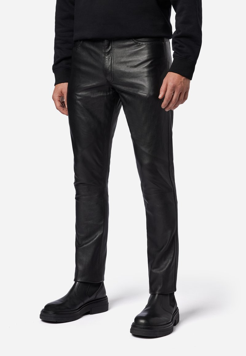 Schwarze Lederhosen mit schmaler Passform, glatter Textur, sichtbaren Nähten und einem mittelhohen Bund, kombiniert mit schwarzen Knöchelboots.